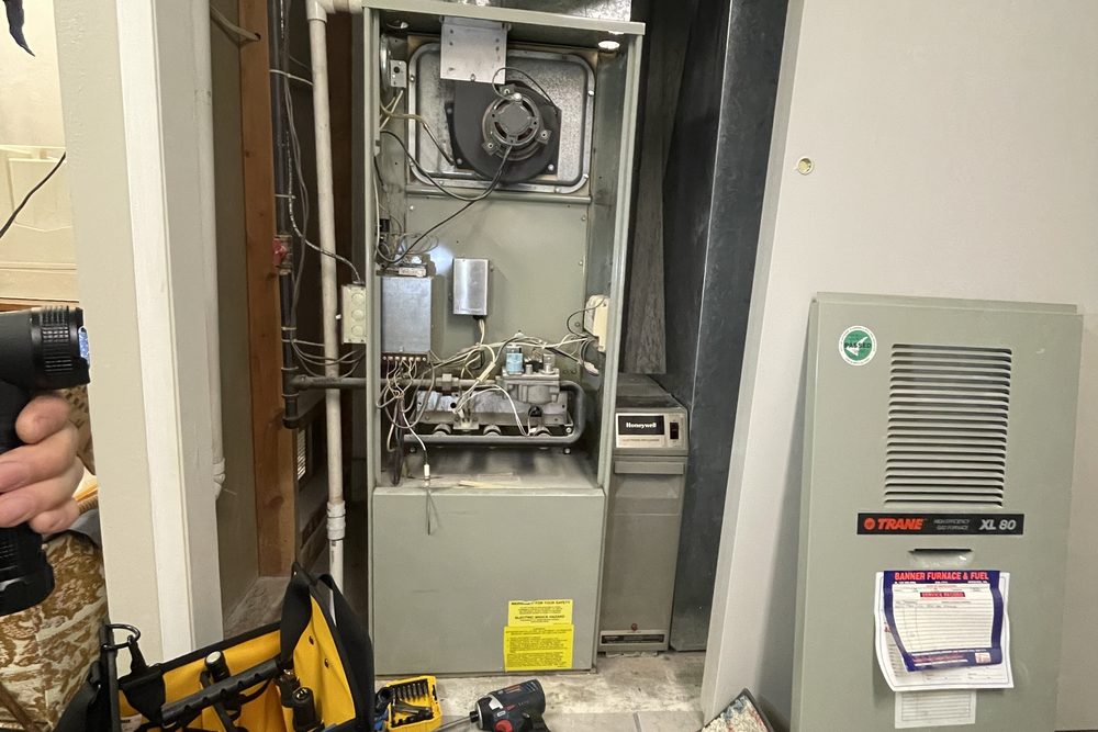Newer furnace beside an older unit in a hallway closet, tools on the floor